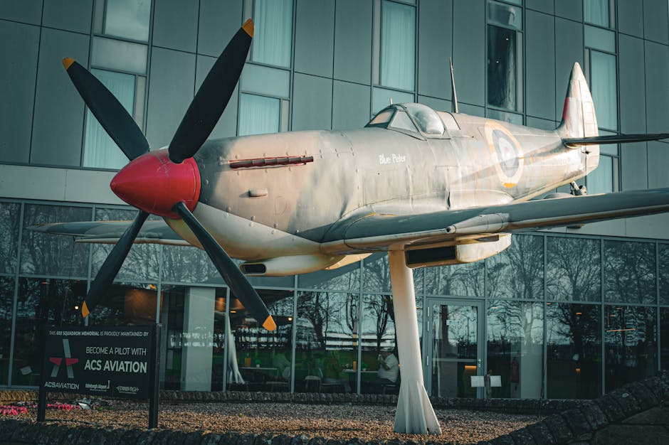 A vintage military fighter plane with a silver-gray fuselage displayed outdoors in front of a modern glass building, reflecting surrounding trees and sky. The aircraft features a bright red nose cone,