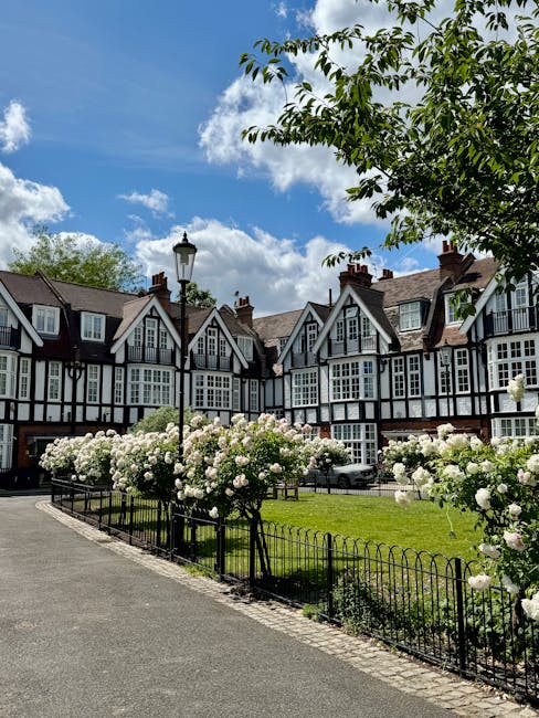 A residential garden area featuring a neatly maintained lawn bordered by a low black metal fence, with blooming white roses along the fence line. Behind the garden, there are classic timber-framed hou