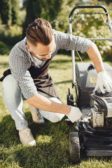A man with brown hair, wearing safety glasses, a checked shirt, and white gloves, is kneeling on a lush green lawn while working on a lawnmower outdoors. The garden features a well-maintained grassy a