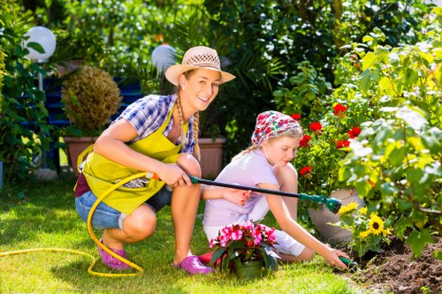 A woman and a young girl are working together in a lush garden with vibrant flowering plants and greenery, which includes a variety of blooming flowers in red, yellow, and pink hues. The woman, dressed in a checkered shirt, denim shorts, a wide-brimmed straw hat, and a bright yellow apron, is crouched down and smiling while assisting the girl, who is wearing a light purple outfit and a colorful sun hat. The girl is using a small gardening trowel to tend to a flower bed, which is bordered by soil and decorative flower arrangements. The garden features a well-maintained lawn with dense foliage and a backdrop of tall trees and shrubs, providing shade and a natural setting. Sunlight filters through the leafy canopy, creating a warm, inviting environment suitable for outdoor gardening activities. Garden tools, including a garden hose, are visible, emphasizing a gardening context, and the scene suggests outdoor maintenance or planting in a well-kept, residential garden, typical of suburban areas in Colindale.